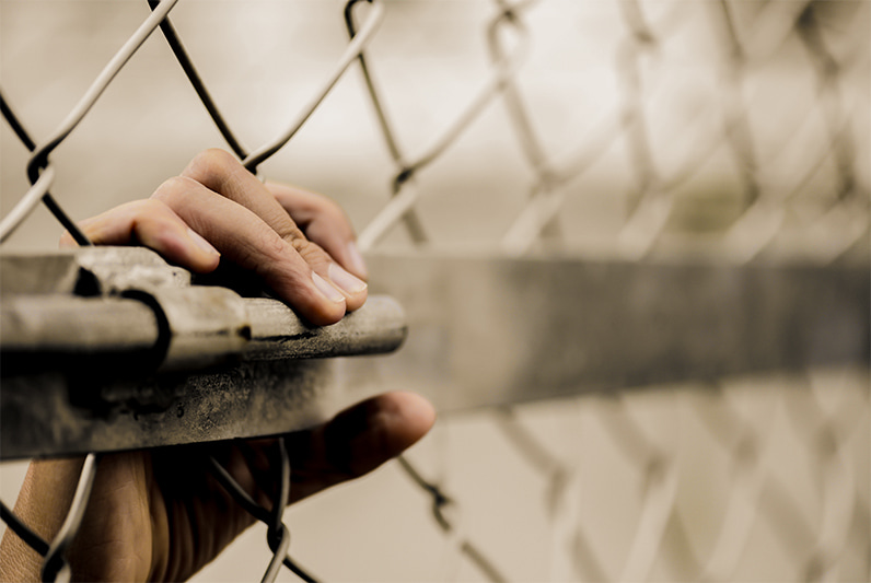 A close-up of a hand gripping a metal chainlink fence from the inside.