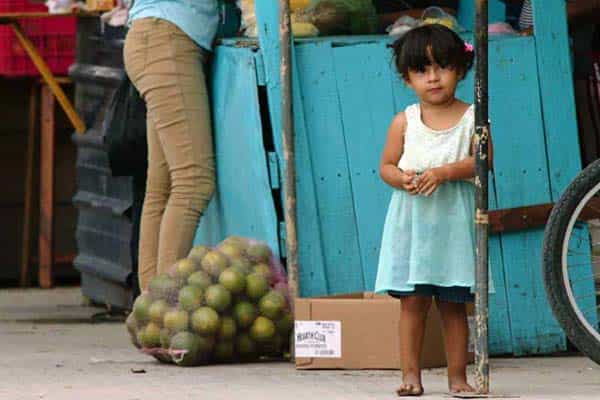 A little girl stands next to a market cart and bag of fruit.
