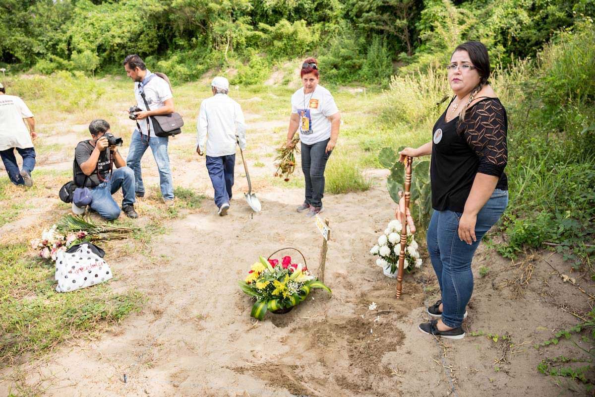 Mothers stand around gravesite with flowers, while photographers take pictures.