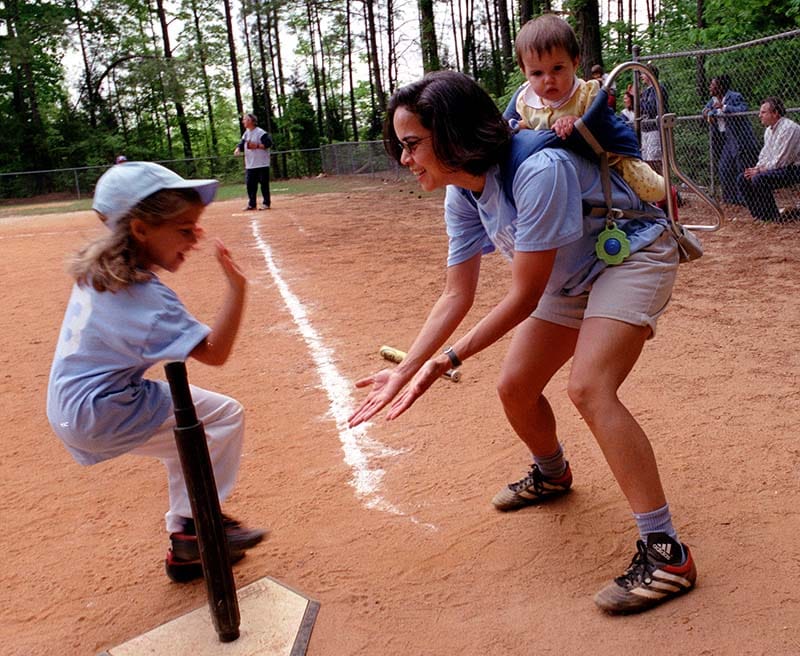 Marie Lynn Miranda with a baby on her back in a carrier, gives high fives to a child. They are wearing softball attire and on a softball field.