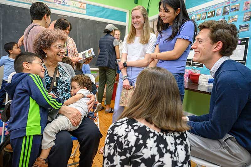 Students talk and smile with families in a classroom. An elderly woman is holding a child and another one leans on her.