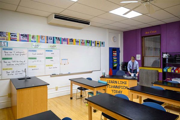 A male teachers stands at his desk in a colorful classroom.