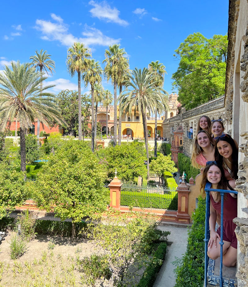 Five female Notre Dame students smile from a balcony window overlooking palm trees and greenery in Alcázar in Sevilla.