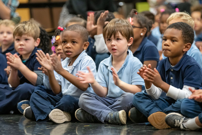 Elementary school children sit cross-legged on the floor while clapping.