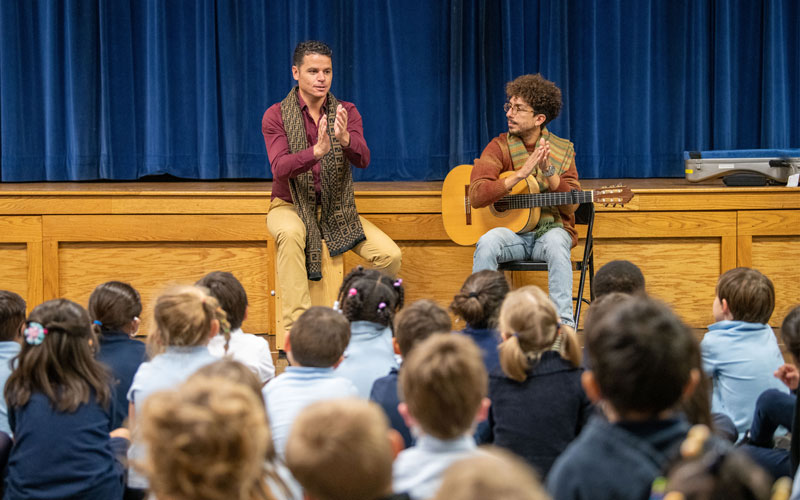 Jaime El Estampio and Antonio Herrera sit side-by-side with their hands clapped together while speaking to a classroom of students.