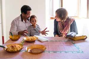 Two adults and a child sit as another adult stands around a table with four bowls full of pasta.