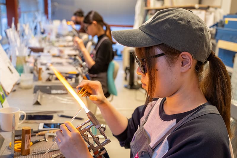 A young woman in a black cap sits at a workstatin. In one hand, she holds a small glass rod over an open flame, in the other, she holds a metal instrument used to quickly cut the hot glass into small beads.