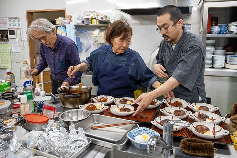 A Japanese woman stands between her son and husband in a large kitchen. She spoons food into bowls for their guests. The counter is cluttered by pots, pans, spices and utensils.
