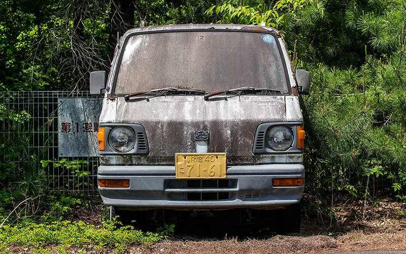 An old, weathered, rusted vehicle sits empty with grass growing around it.
