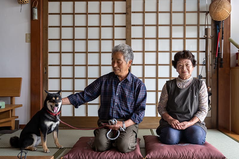 Retired rice farmers Seiko and Masao Sato sit on their knees on cushions with their Shiba Inu dog on a leash beside them.