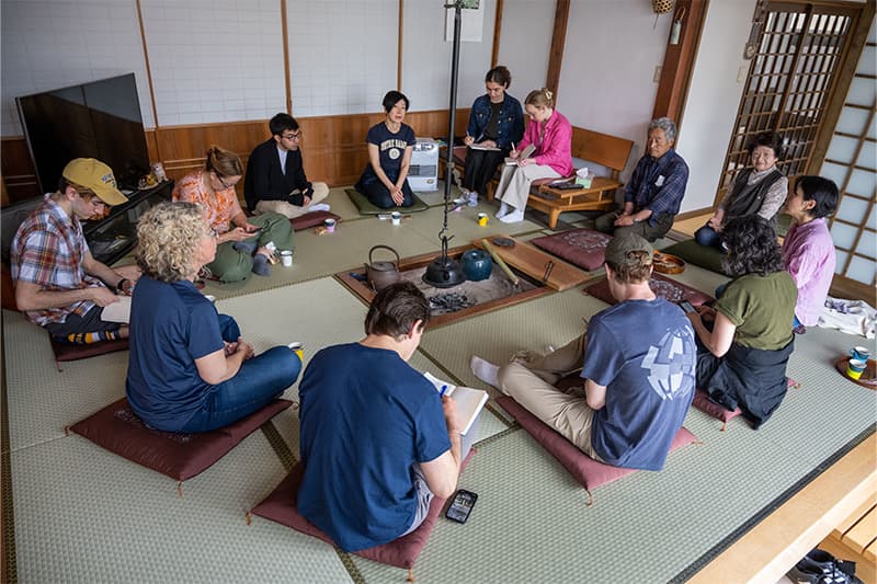The Notre Dame research team sit on cushions around a traditional Japanese irori, a sunken hearth with a tea kettle hanging from the ceiling, taking notes. They are joined by retired rice farmers Seiko and Masao Sato.