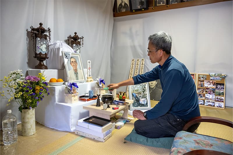 A Japanese man sits on his knees in front of a makeshift alter. He lights a candle in front of a framed image of his father.