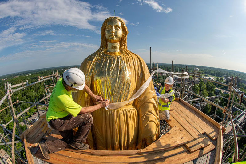 Two people stand on scaffolding next to the statue of Mary on top of the Golden Dome. They're applying gold leaf, and the paper the leaf is on is now empty and blowing in the breeze.