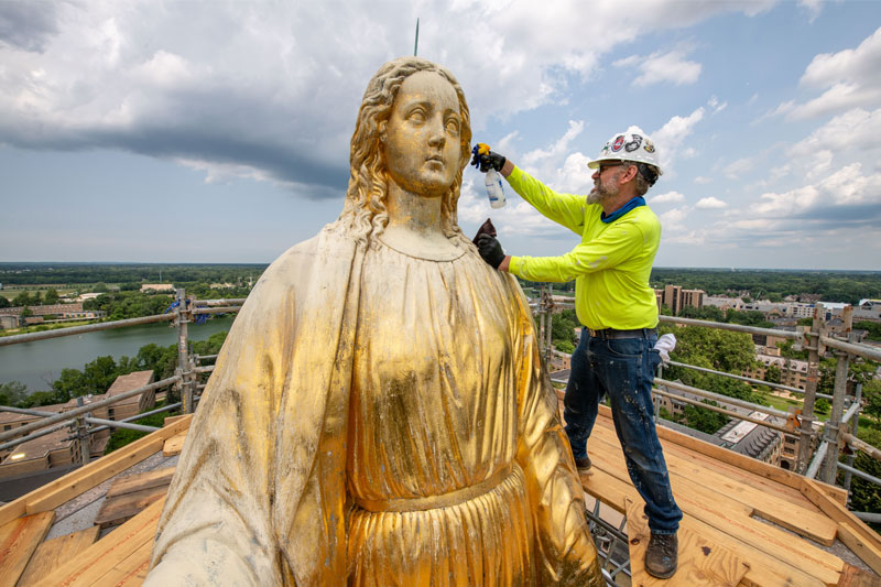 A man is standing on scaffolding next to the statue of Mary on the Golden Dome. He's spraying the surface of the statue with a squirt bottle.
