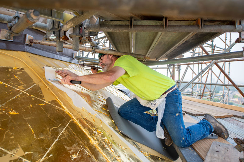 A man kneels on scaffolding while applying gold leaf to the Golden Dome.
