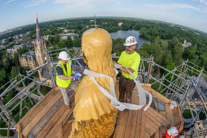 Two men stand at the statue of Mary on top of the Golden Dome. A thin strip of paper surrounds her.
