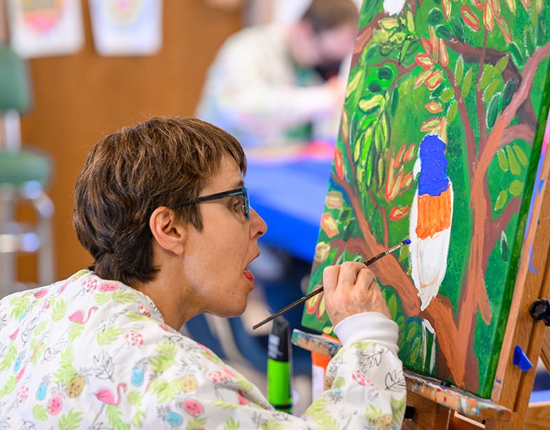 A resident of Misericordia is closely working on a painting with paint brush in hand.