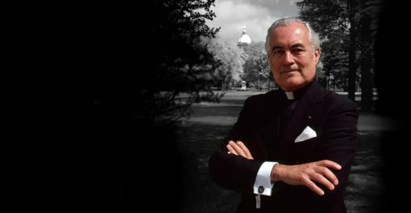 Father Ted Hesburgh is pictured standing against a black backdrop.