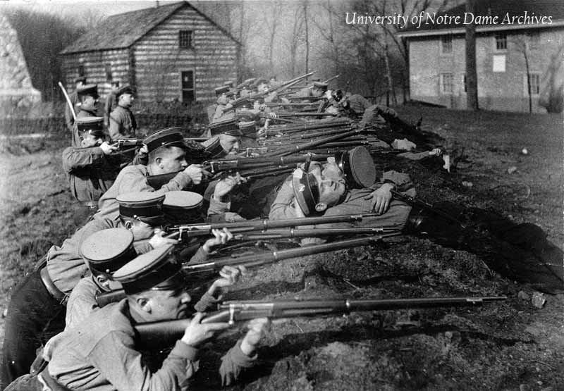 Students in the military training corps at rifle target practice in the trenches, 1915-1916. The Founder's Monument, Log Chapel, and Old College are in the background.