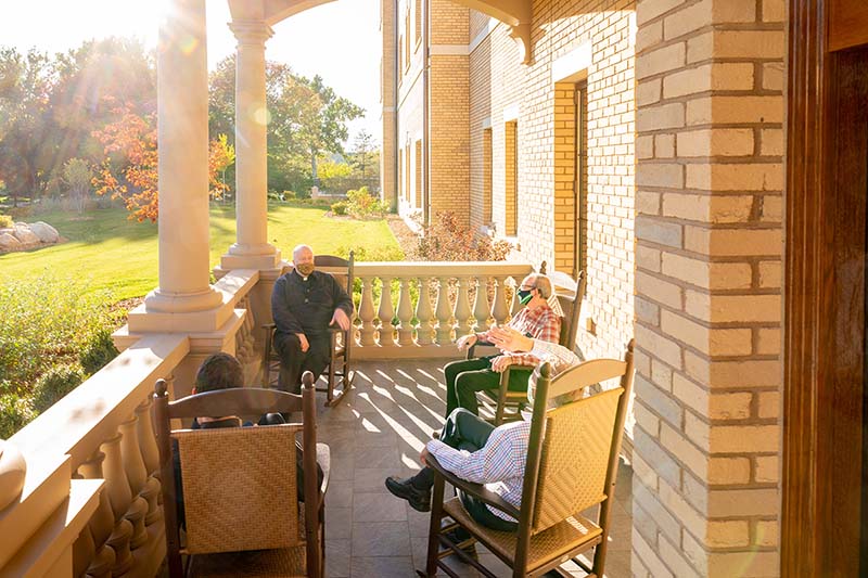 Four men wearing masks sit on the front porch of Corby Hall on a sunny evening.