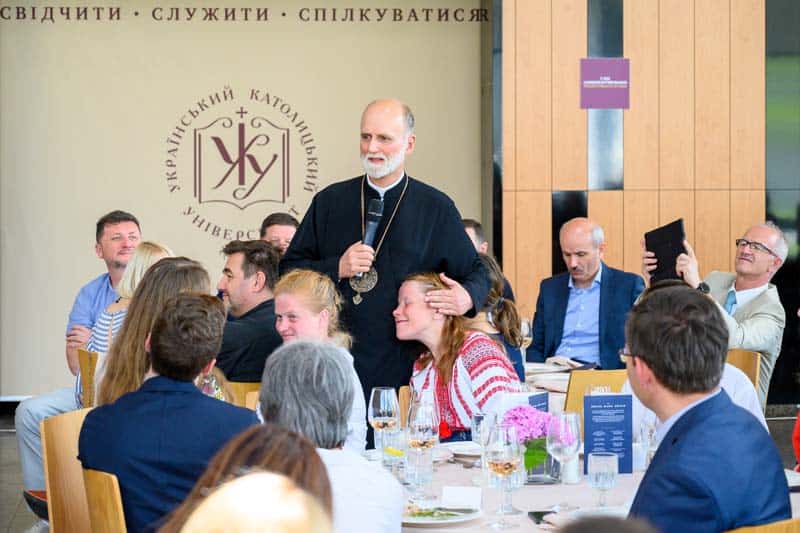 The Right Reverend Borys Gudziak, Archeparch of Philadelphia holds a microphone giving remarks at a ceremony while hugging one of the residents.