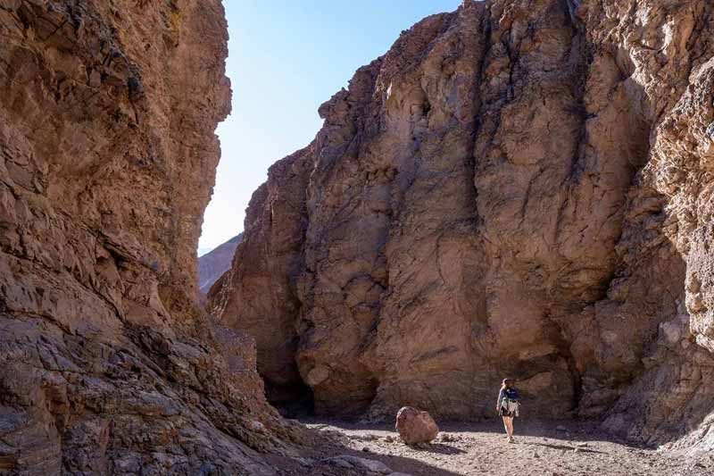 A college student wearing a backpack walks between high walls of red rock in Death Valley, with a boulder beside her.