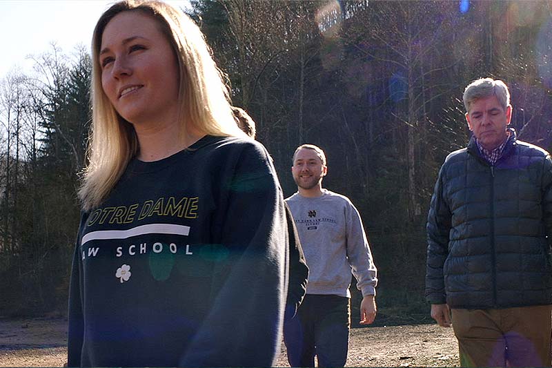 A female student wearing a Notre Dame Law School sweatshirt walking in a wooded field. Three other individuals walk behind her.