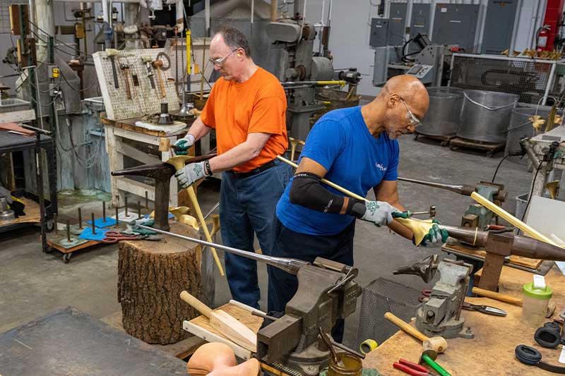 Two men in safety goggles and gloves make trumpet bells in a manufacturing plant.