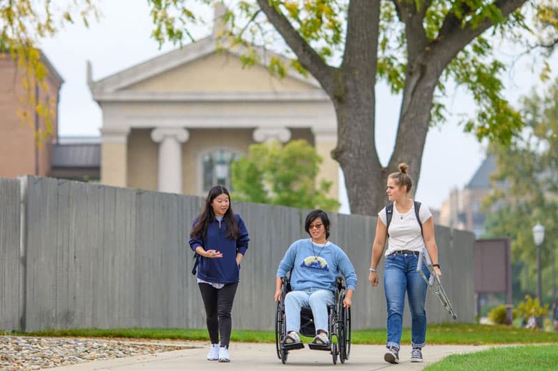 A student using a wheelchair rides down the sidewalk between two people, one who is carrying crutches.