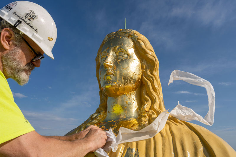 A man in a hard hat applies gold leaf to the statue of Mary on top of the Golden Dome.