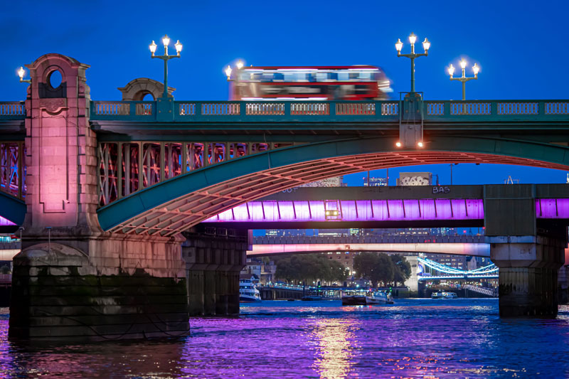 A image of a blurry red double-decker buse as it travels on a bridge over the River Thames at night.