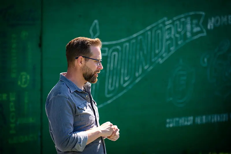 Clint Carlson speaking in front of a green painted wall with a 'Foundry Field' pennant shape.
