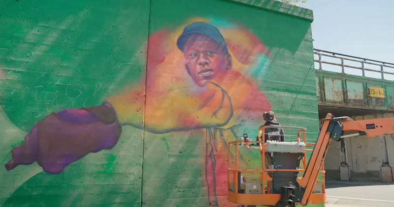 A photo of a man in a hat standing on a lift, using spray paint to paint a mural.