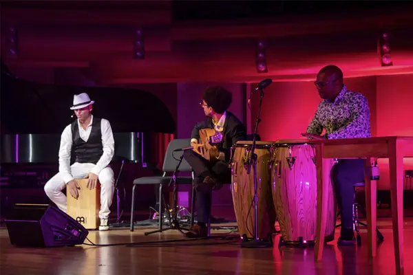 Jaime El Estampio sits on a wooden drum while Antonio Herrera plays guitar and an artist beats on hand drums.