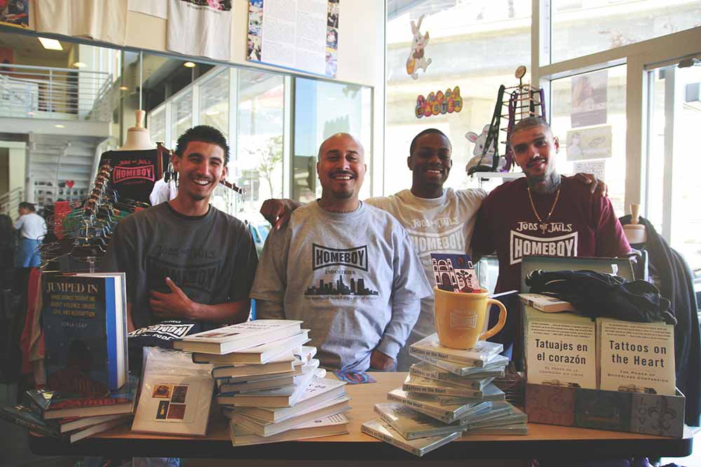 Four men wearing Homeboy shirts standing behind a stack of books