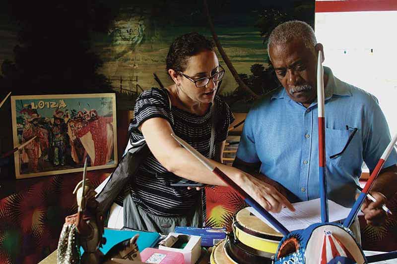 A woman and a man look at notes on pieces of paper. The woman is pointing at the middle of the paper. There are objects and figures on the desk next to them.