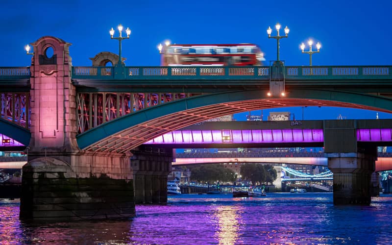 A image of a blurry red double-decker buse as it travels on a bridge over the River Thames at night. 