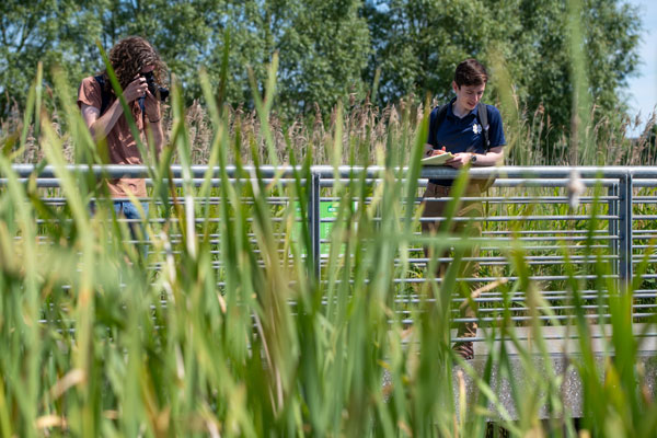 Two young men, one with a camera and the other with a notebook, stand on a bridge. They're overlooking the water at Fr. Collins Park in Ireland.