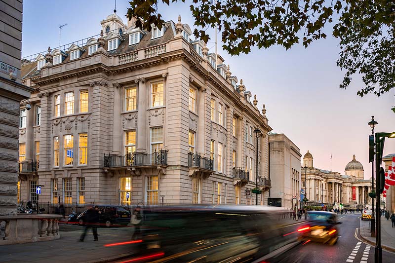 Cars drive past a stone building with big, well-lit windows.