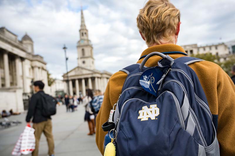 A student with his back to us dons a Notre Dame backpack. Historical buildings are in front of him.