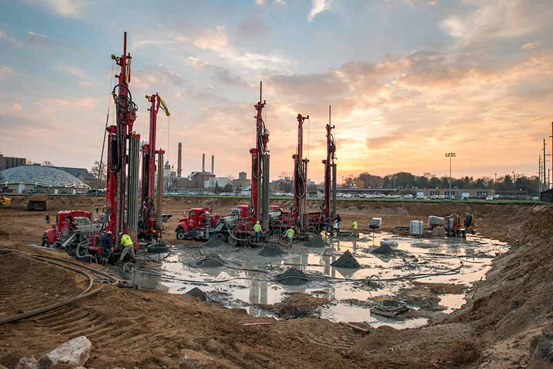 Five well drilling trucks parked on a dirt construction site.