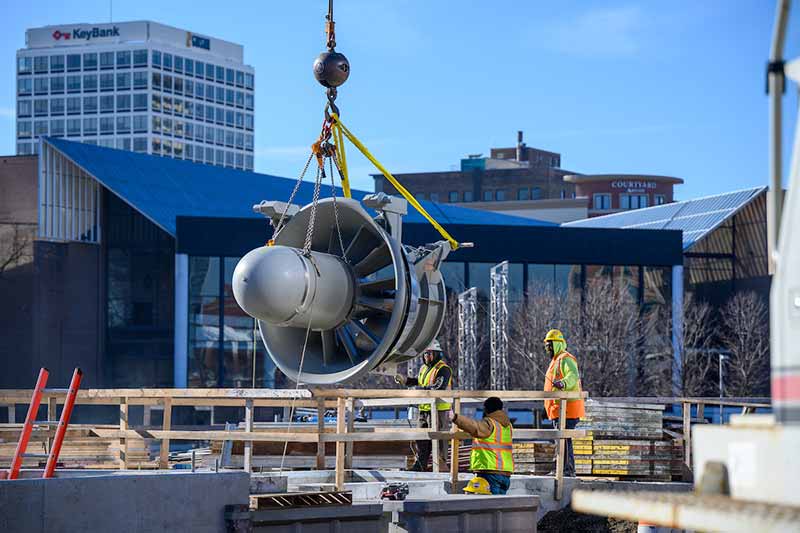Three construction workers wearing protective gear help with a large turbine on a construction site in a downtown area.