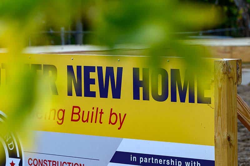 A yellow sign that reads New Home, with tree branches in the foreground.