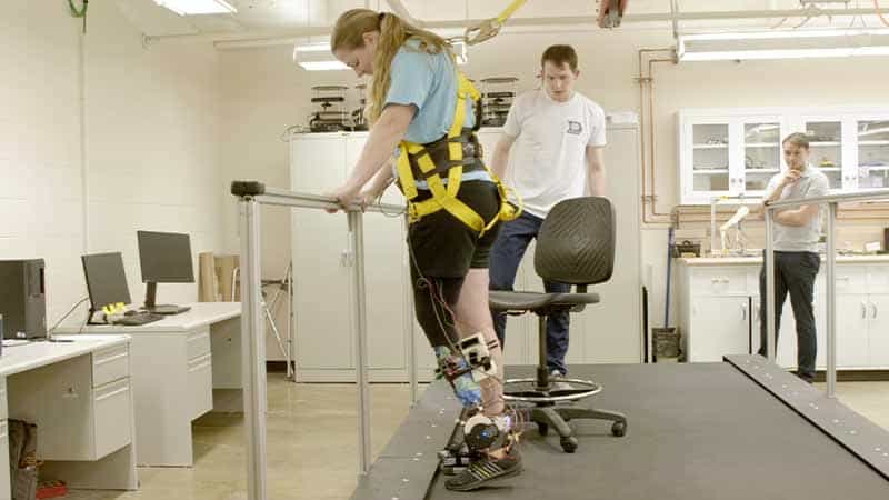A woman wearing a bright yellow harness works on standing on her tiptoes while testing out a powered prosthetic lower-leg.