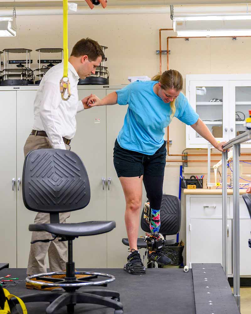 A man helps a woman wearing a powered prosthetic lower-leg onto a treadmill inside of a lab.