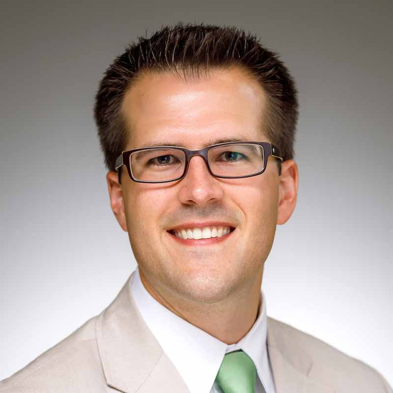 A headshot of Patrick Wensing, a man wearing glasses, a light gray suit, and lime green tie. 