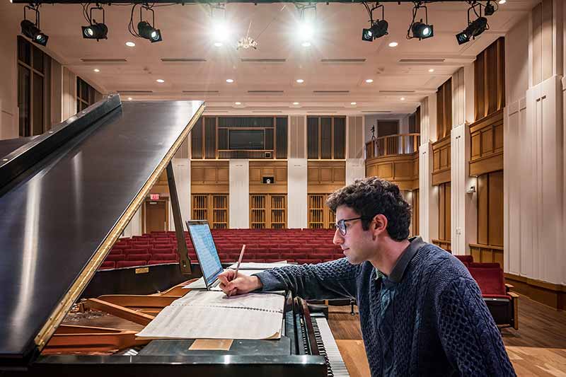 A man sits on a piano bench on a stage inside of a theatre and writes on a piece of paper with a pencil.