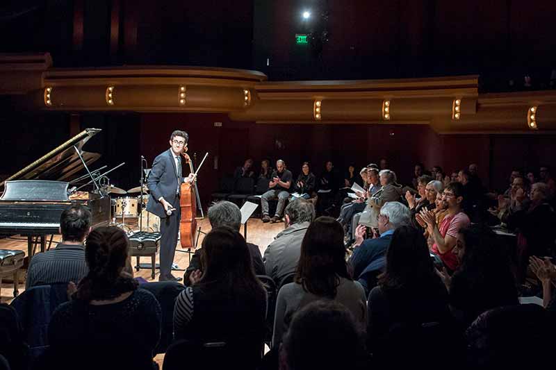 A man, holding a cello, standing next to a piano on stage, looks out to the cheering audiance.