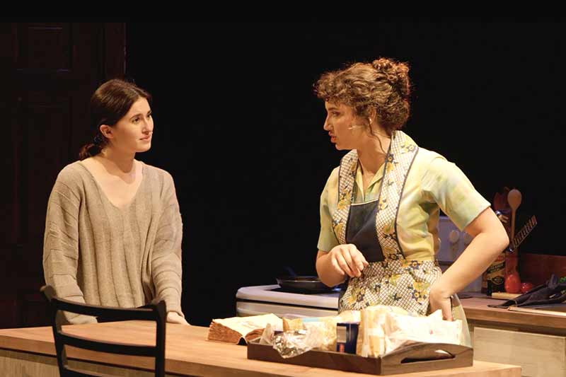 Two women on a stage, one wearing a sweater and the other an apron, stand next to a table with italian pasta placed next to them.