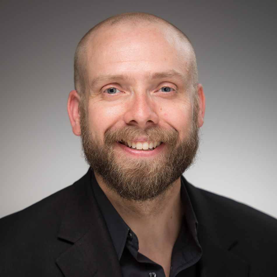 A headshot of a bearded, bald man against a light gray background, wearing a black shirt.